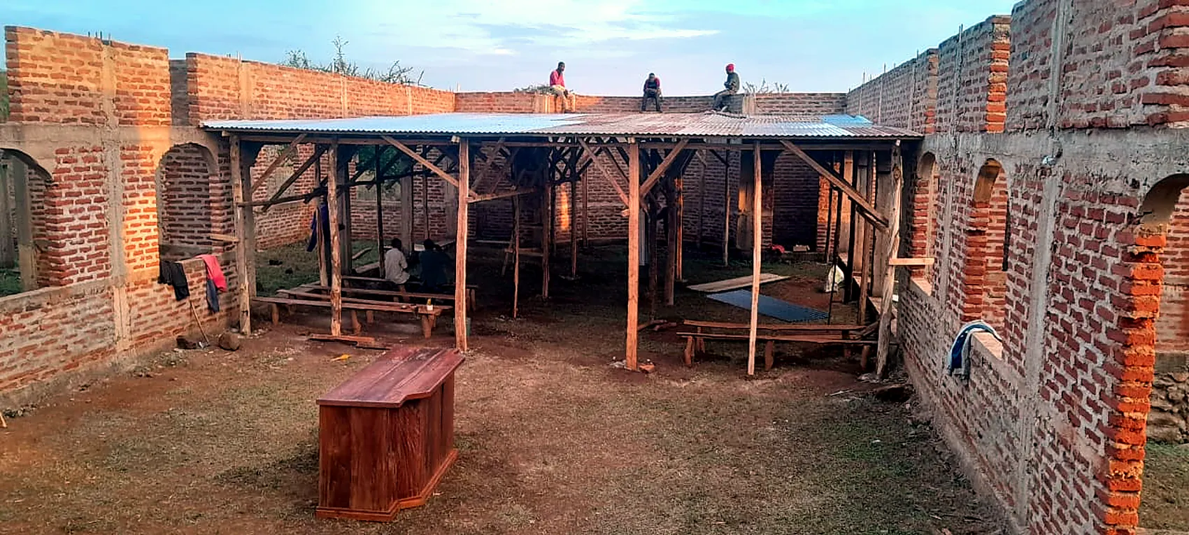 Dusk inside the new church. A temporary roof of poles and metal sheeting covers part of the span, and three men sit on the top of the finished wall.