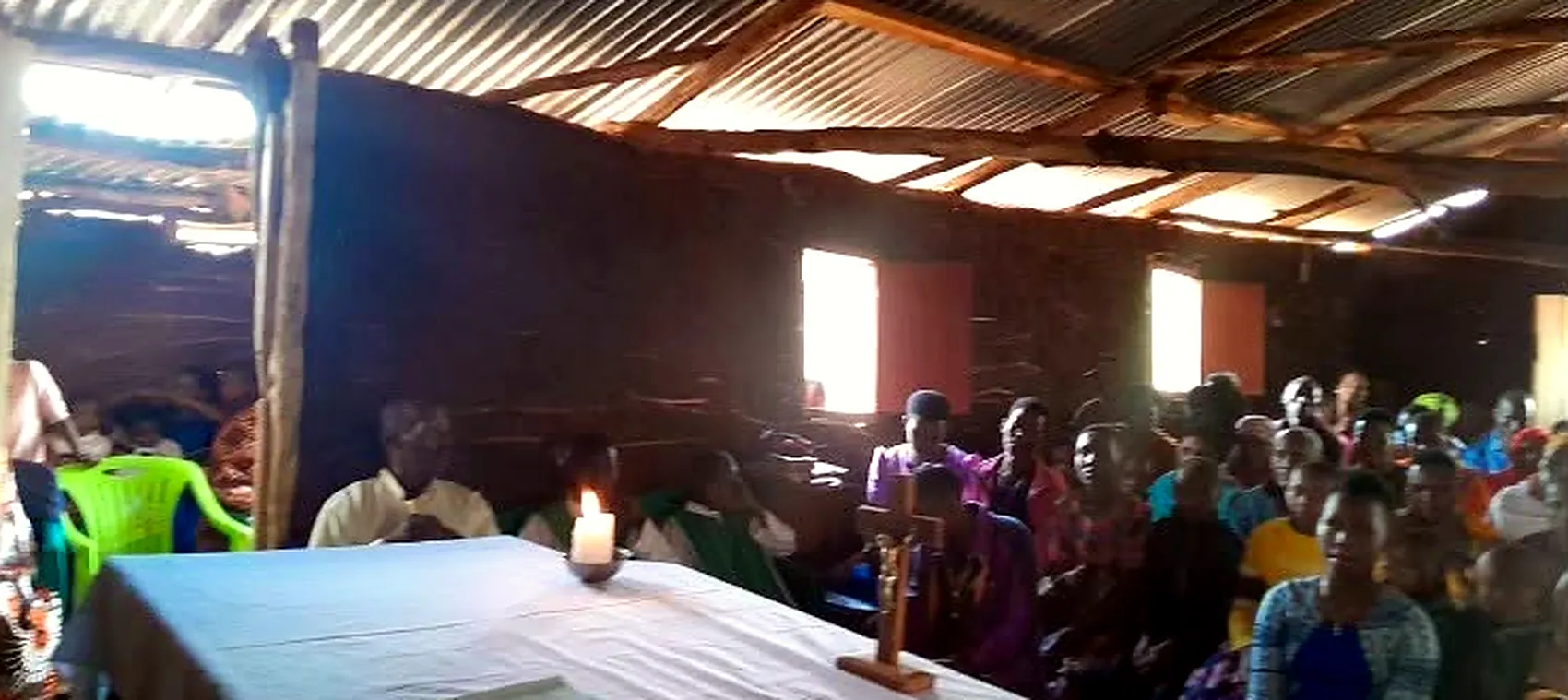 Earth walls, a corrugated roof, and a candle lit on the altar table in the middle of the day. The light comes from two openings cut in the wall.