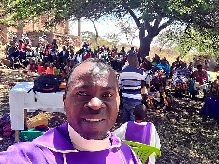 Father Vicent Mbusa, CSC, in purple vestments, his congregation gathered under the trees behind him.